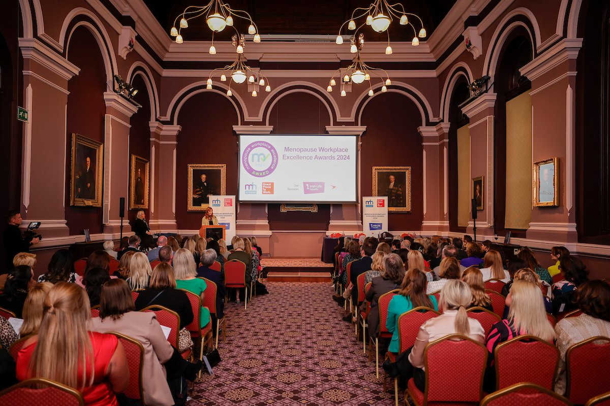 A full auditorium with chandeliers and red seating facing a stage carrying Menopause Workplace Excellence Awards branding