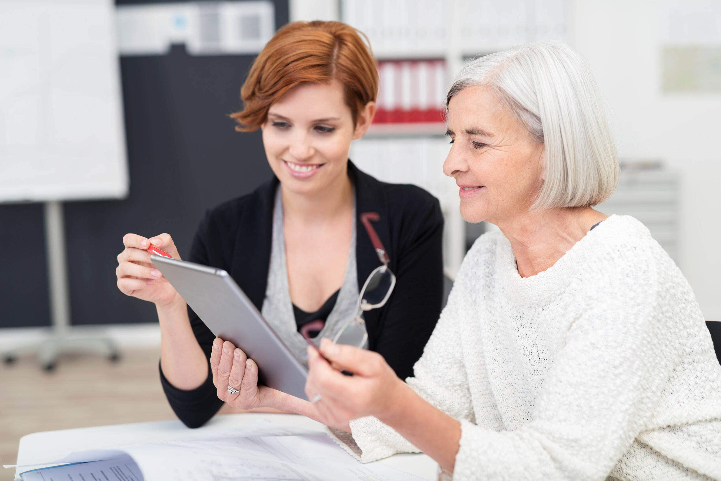 Two women sitting either side of a desk looking together at a tablet, with a pink flower out of focus in the foreground